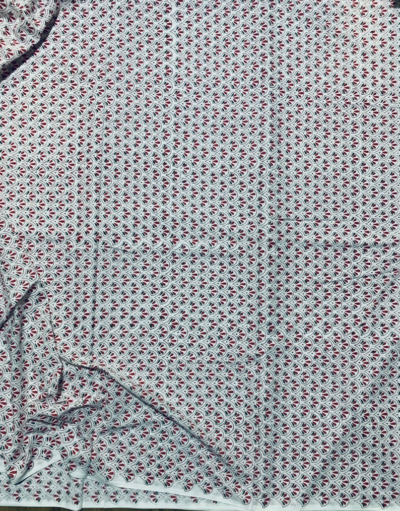 May include: White fabric with a repeating pattern of red and black fan-shaped designs. The fabric is folded, showing the texture and drape of the material. The pattern is evenly spaced across the surface.