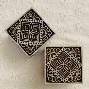 May include: Two wooden printing blocks with intricate floral and geometric designs. One block is diamond-shaped, the other square. The blocks are dark brown with white accents, set against a white, textured background.