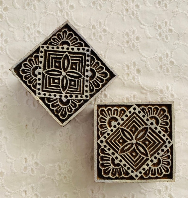May include: Two wooden printing blocks with intricate floral and geometric designs. One block is diamond-shaped, the other square. The blocks are dark brown with white accents, set against a white, textured background.