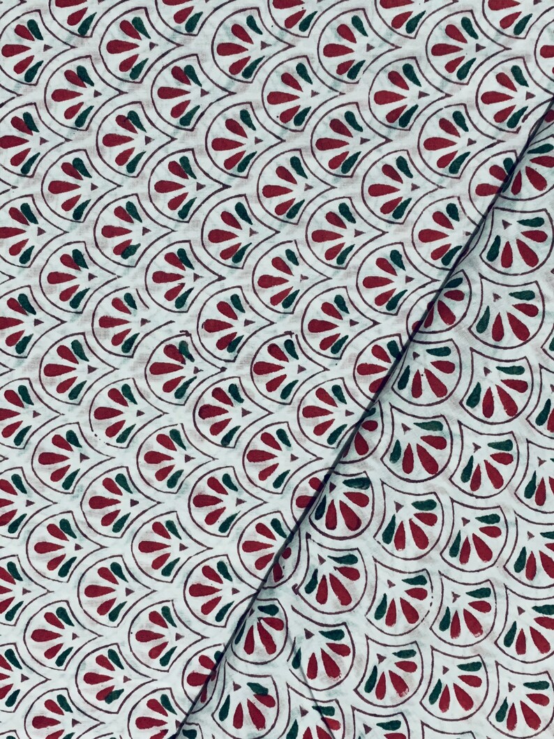 May include: White fabric with a repeating pattern of stylized, fan-shaped designs in dark red and green. The pattern is outlined in a dark brown color. The fabric is folded diagonally, showing the pattern's continuity.