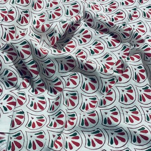 May include: White fabric with a repeating pattern of stylized red and green floral shapes. The fabric is draped, showing folds and shadows that add depth to the design. The pattern is evenly spaced across the material.