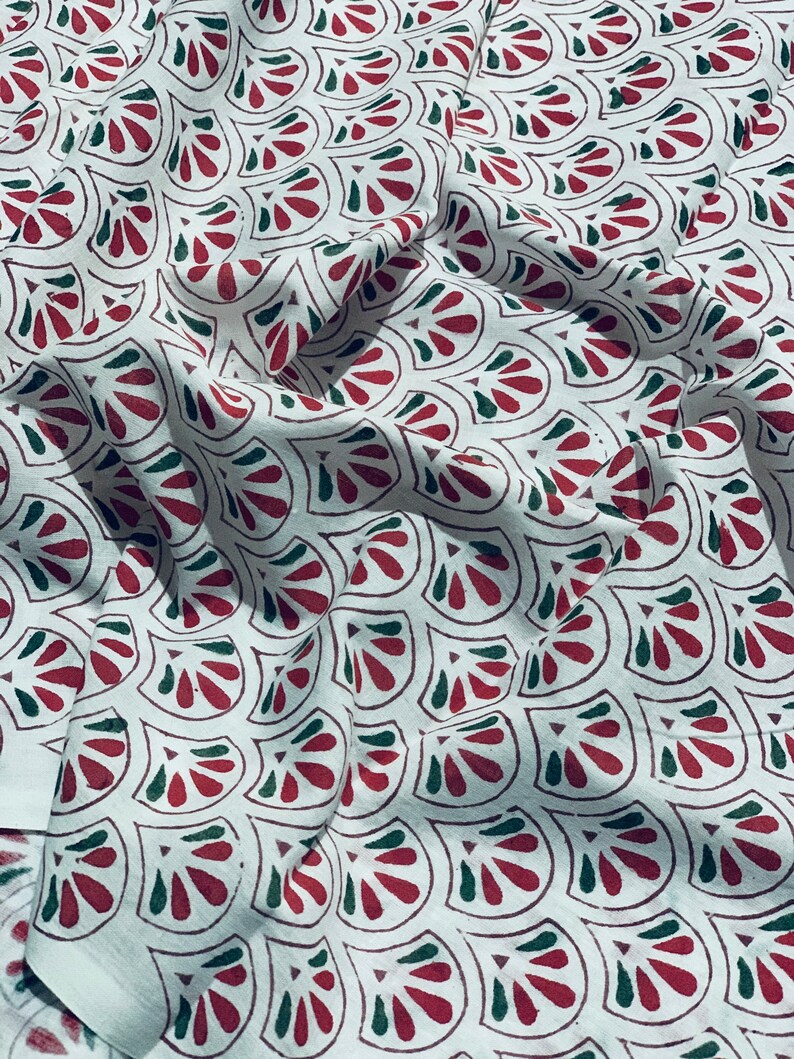 May include: White fabric with a repeating pattern of stylized red and green floral shapes. The fabric is draped, showing folds and shadows that add depth to the design. The pattern is evenly spaced across the material.