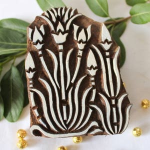May include: A wooden block printing stamp with a white floral design. The design features stylized flowers with long stems and leaves. The stamp is carved from dark wood and has a natural, rustic look.