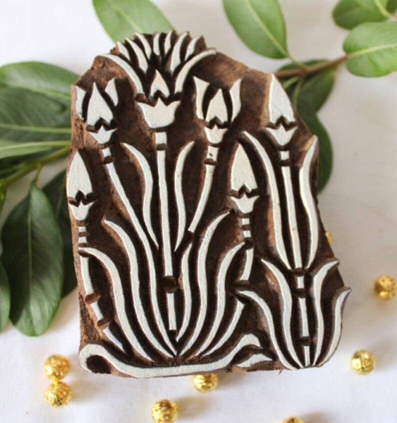 May include: A wooden block printing stamp with a white floral design. The design features stylized flowers with long stems and leaves. The stamp is carved from dark wood and has a natural, rustic look.