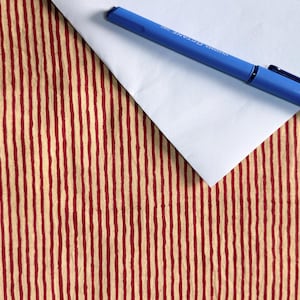 May include: A blue pen rests on a white envelope, set against a backdrop of a red and beige striped pattern. The pen is a solid blue color, and the envelope is plain white. The background has vertical stripes.