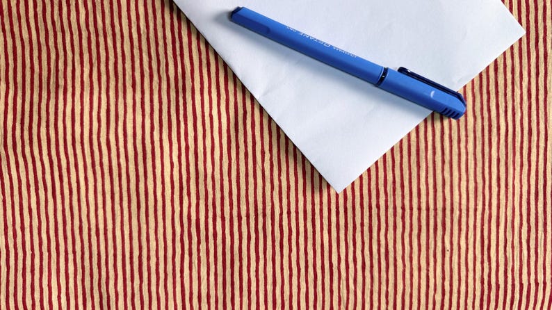 May include: A blue pen rests on a white envelope, set against a backdrop of a red and beige striped pattern. The pen is a solid blue color, and the envelope is plain white. The background has vertical stripes.
