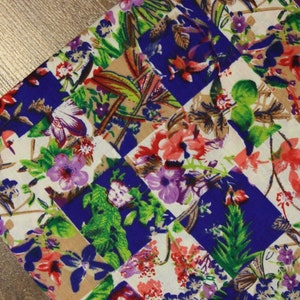 May include: A colourful fabric with a patchwork design featuring a variety of flowers and leaves in shades of pink, purple, green, and blue. The fabric has a checkered pattern with alternating squares of beige and blue.