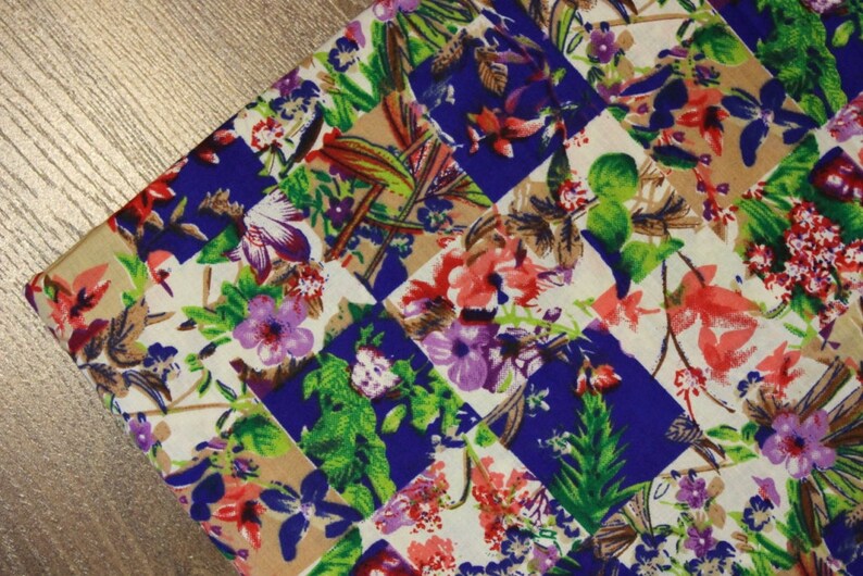 May include: A colourful fabric with a patchwork design featuring a variety of flowers and leaves in shades of pink, purple, green, and blue. The fabric has a checkered pattern with alternating squares of beige and blue.