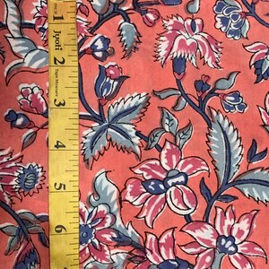 May include: A close-up of a pink fabric with a floral pattern. The fabric has a repeating pattern of blue and pink flowers with green leaves. A yellow tape measure is visible on the left side of the image.