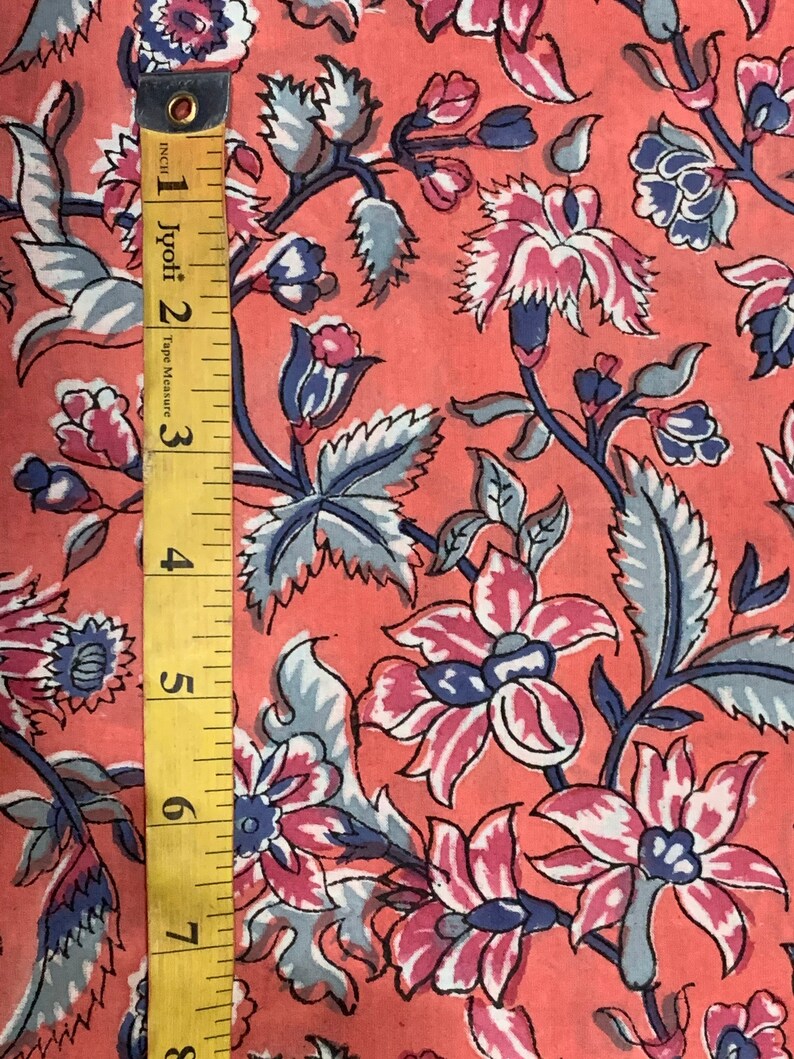 May include: A close-up of a pink fabric with a floral pattern. The fabric has a repeating pattern of blue and pink flowers with green leaves. A yellow tape measure is visible on the left side of the image.