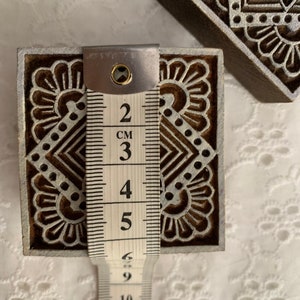 May include: Wooden block printing stamps with intricate floral and geometric designs. The stamp measures approximately 2 inches (5 cm) in height. The stamps are brown with white detailing. The image shows the stamp being measured with a measuring tape.