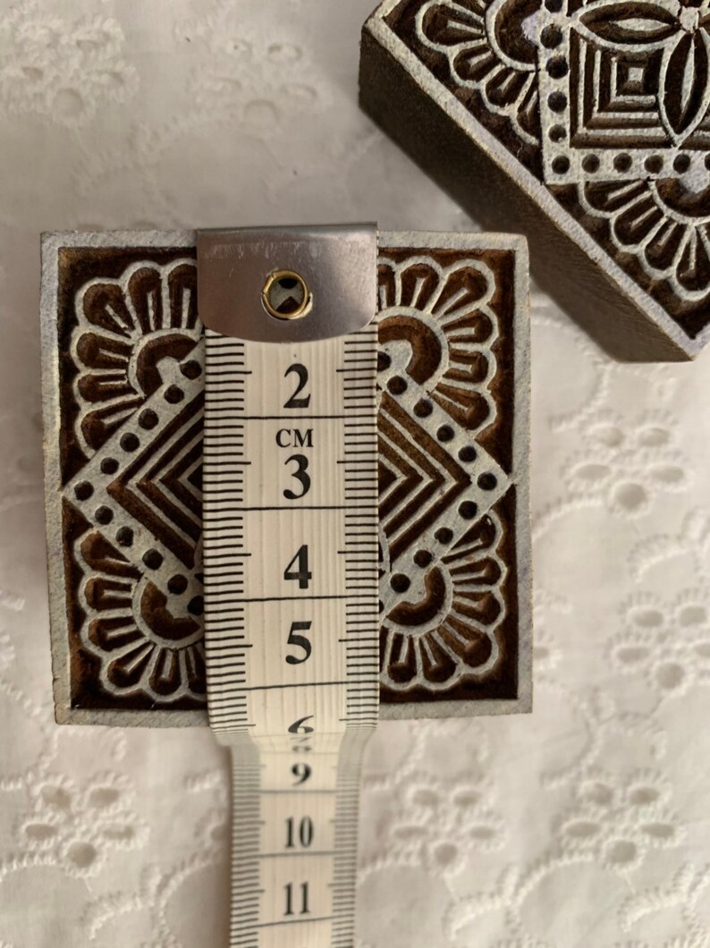 May include: Wooden block printing stamps with intricate floral and geometric designs. The stamp measures approximately 2 inches (5 cm) in height. The stamps are brown with white detailing. The image shows the stamp being measured with a measuring tape.