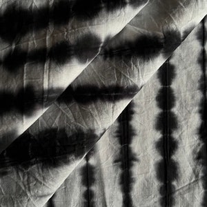 Puede incluir: Primer plano de una tela con un patrón de teñido anudado. La tela es blanca con rayas verticales de teñido anudado negro. La tela está doblada sobre sí misma, creando líneas diagonales y sombras. La tela parece ser de algodón o un material similar.