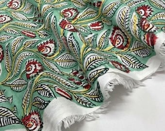 Op de afbeelding: Een stuk stof met een lichtgroene achtergrond en een herhalend patroon van rode en witte bloemen en gele bladeren. Het textiel heeft een witte rand en een gerafelde, onafgewerkte rand aan de onderkant van het materiaal.