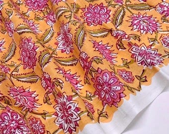 May include: A piece of fabric featuring a vibrant orange background with a repeating floral pattern in shades of pink, white, and olive green. The textile displays a distinct white border along the edge, showcasing a detailed block-printed design.