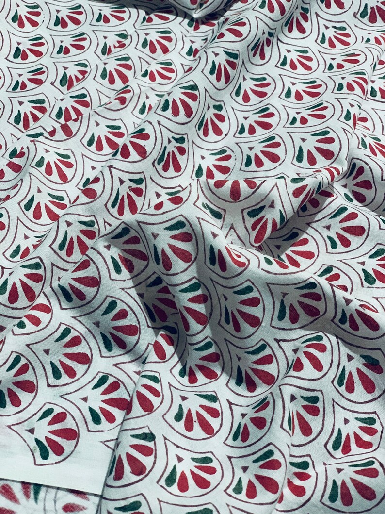 May include: White fabric with a repeating pattern of stylized red and green fan-like shapes. The fabric is slightly wrinkled, suggesting it is a soft material, and the pattern is evenly distributed across the surface.