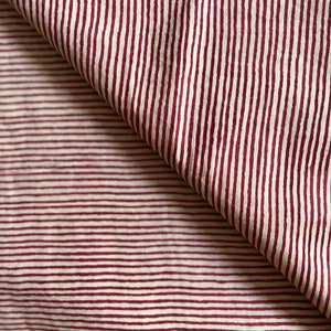 May include: Close-up of a fabric with a repeating pattern of burgundy vertical stripes on an off-white background. The fabric is folded over, revealing the pattern's continuity. The stripes are evenly spaced and create a textured appearance.