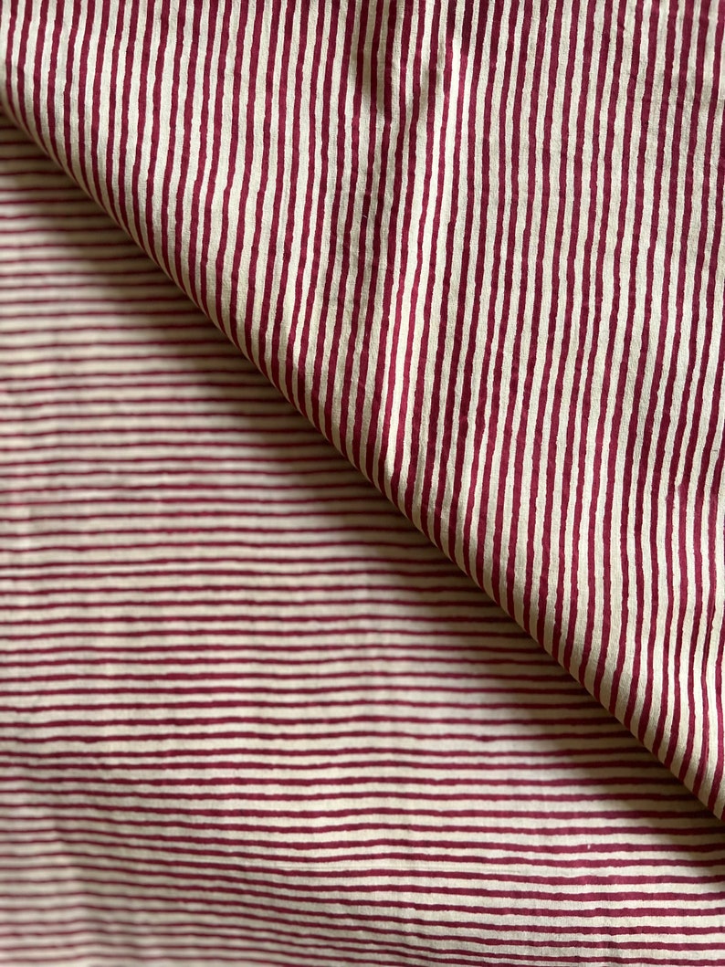May include: Close-up of a fabric with a repeating pattern of burgundy vertical stripes on an off-white background. The fabric is folded over, revealing the pattern's continuity. The stripes are evenly spaced and create a textured appearance.