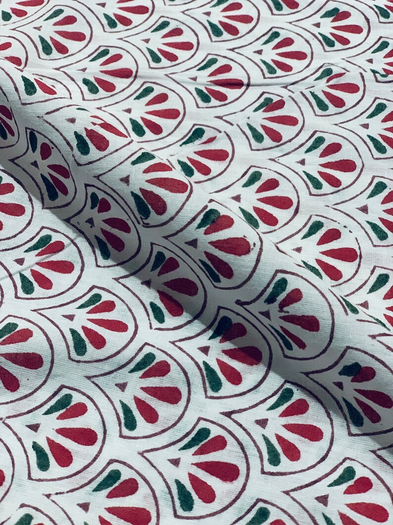May include: White fabric with a repeating pattern of red and green stylized leaf shapes. The leaves are outlined in a dark red-brown color. The fabric is folded, showing the texture and drape of the material.