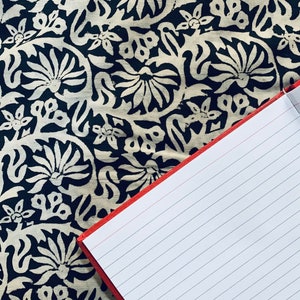 May include: A red notebook with lined pages is lying open on a black and white fabric with a floral pattern.
