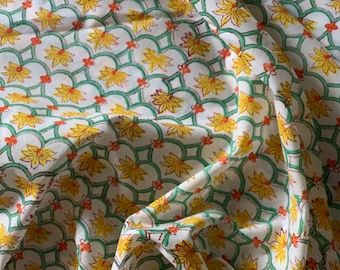 Op de afbeelding: Witte stof met een herhalend patroon van gele bloemen, groene bladeren en oranje accenten. De stof is gevouwen en gedrapeerd, waardoor het patroon zichtbaar is.
