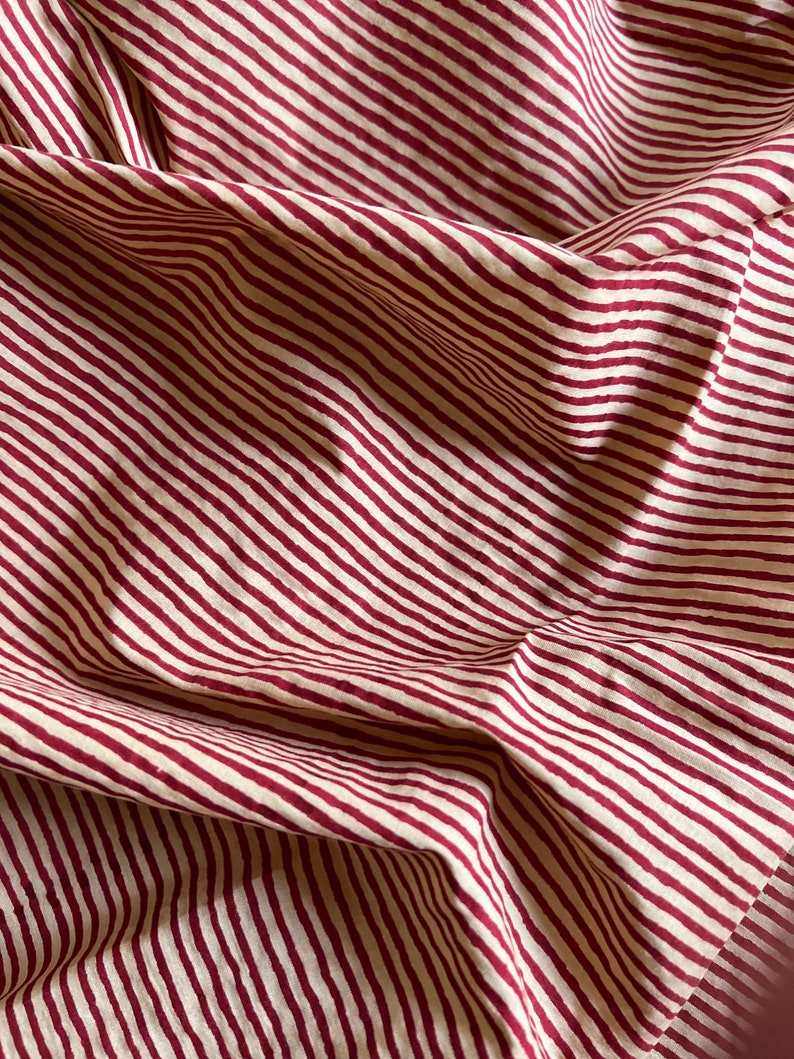 May include: Close-up of a fabric with a repeating pattern of burgundy and cream-colored diagonal stripes. The fabric is folded, creating shadows and texture. The material appears to be cotton or linen.