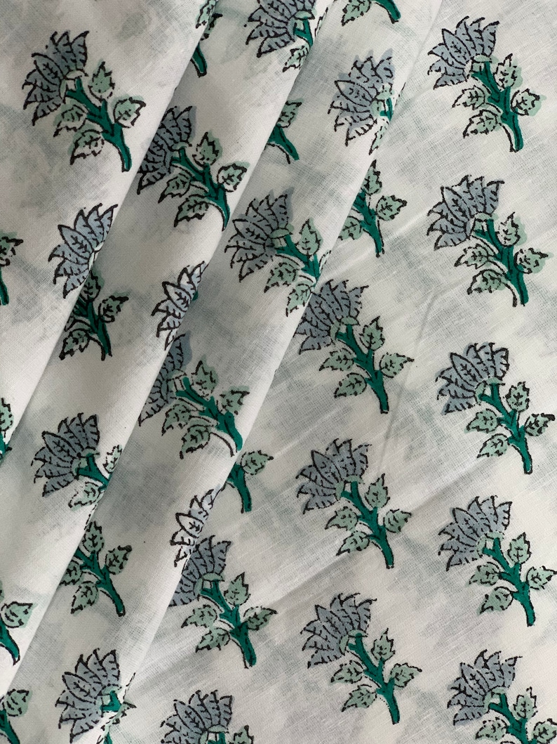 May include: White cotton fabric with a repeating pattern of blue and green floral designs. The fabric is folded in a few places, showing the pattern from different angles.