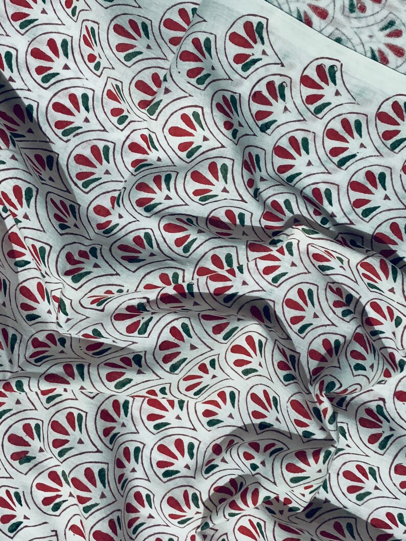 May include: White fabric with a repeating pattern of stylized red and green fan-shaped designs. The fabric has a slightly wrinkled texture, suggesting it is a soft material like cotton or linen. The pattern is outlined in a dark brown color.