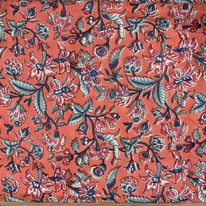 May include: A coral pink fabric with a repeating floral pattern. The flowers are in shades of pink, white, and blue, and the leaves are a light blue-green.