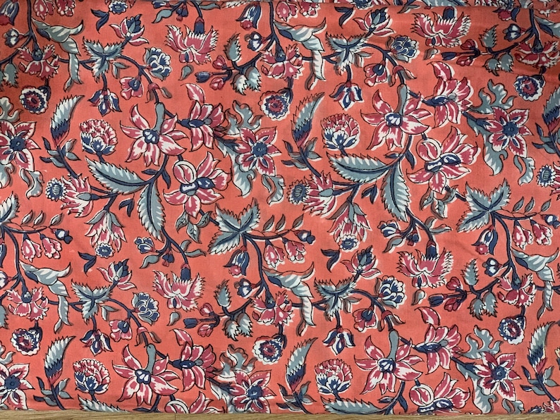 May include: A coral pink fabric with a repeating floral pattern. The flowers are in shades of pink, white, and blue, and the leaves are a light blue-green.