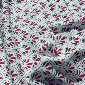 May include: White fabric with a repeating pattern of red and green stylized floral shapes. The fabric is folded, creating shadows and texture. The design is printed on the fabric.
