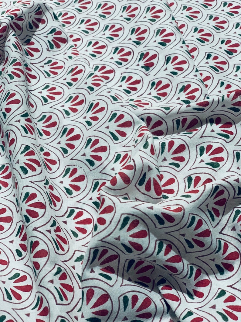 May include: White fabric with a repeating pattern of red and green stylized floral shapes. The fabric is folded, creating shadows and texture. The design is printed on the fabric.