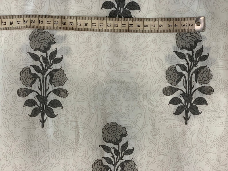 May include: White fabric with a repeating pattern of black floral designs. The fabric is measured with a ruler that shows inches.