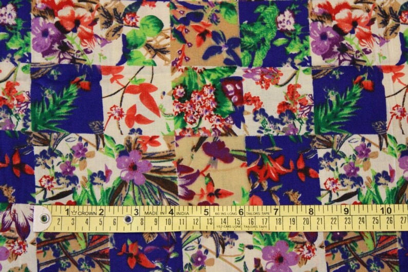 May include: A patchwork fabric with a floral pattern in shades of red, green, purple, and white on a blue and beige background. The fabric is measured with a yellow tape measure.