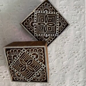May include: Two wooden printing blocks with intricate, symmetrical designs. The blocks feature a square pattern with floral and geometric elements, perfect for textile or paper crafts. The wood has a natural brown color, and the design is in white.
