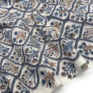 Op de afbeelding: Een witte stof met een herhalend ruitpatroon omlijnd in donkergrijs. Elke ruit bevat een bloemmotief in tinten van gedempt blauw en lichtbruin, wat zorgt voor een consistent botanisch ontwerp over het gehele textieloppervlak.