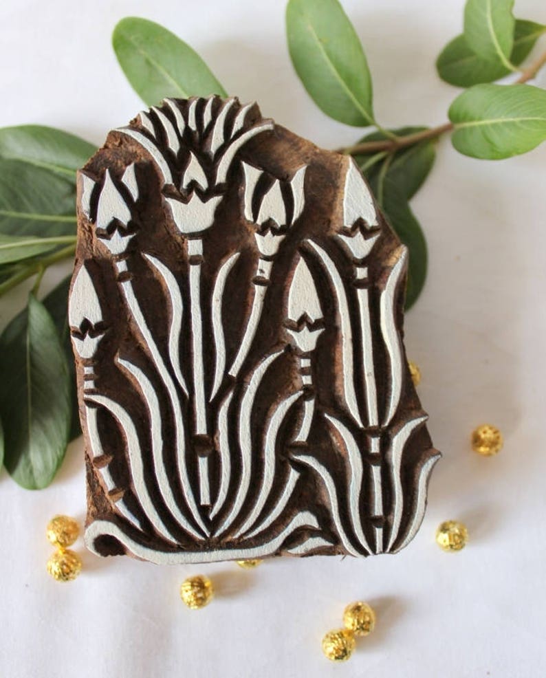 May include: A wooden printing block with a white floral design. The design features five stylized flowers with long stems and leaves. The block is brown and has a rough texture.