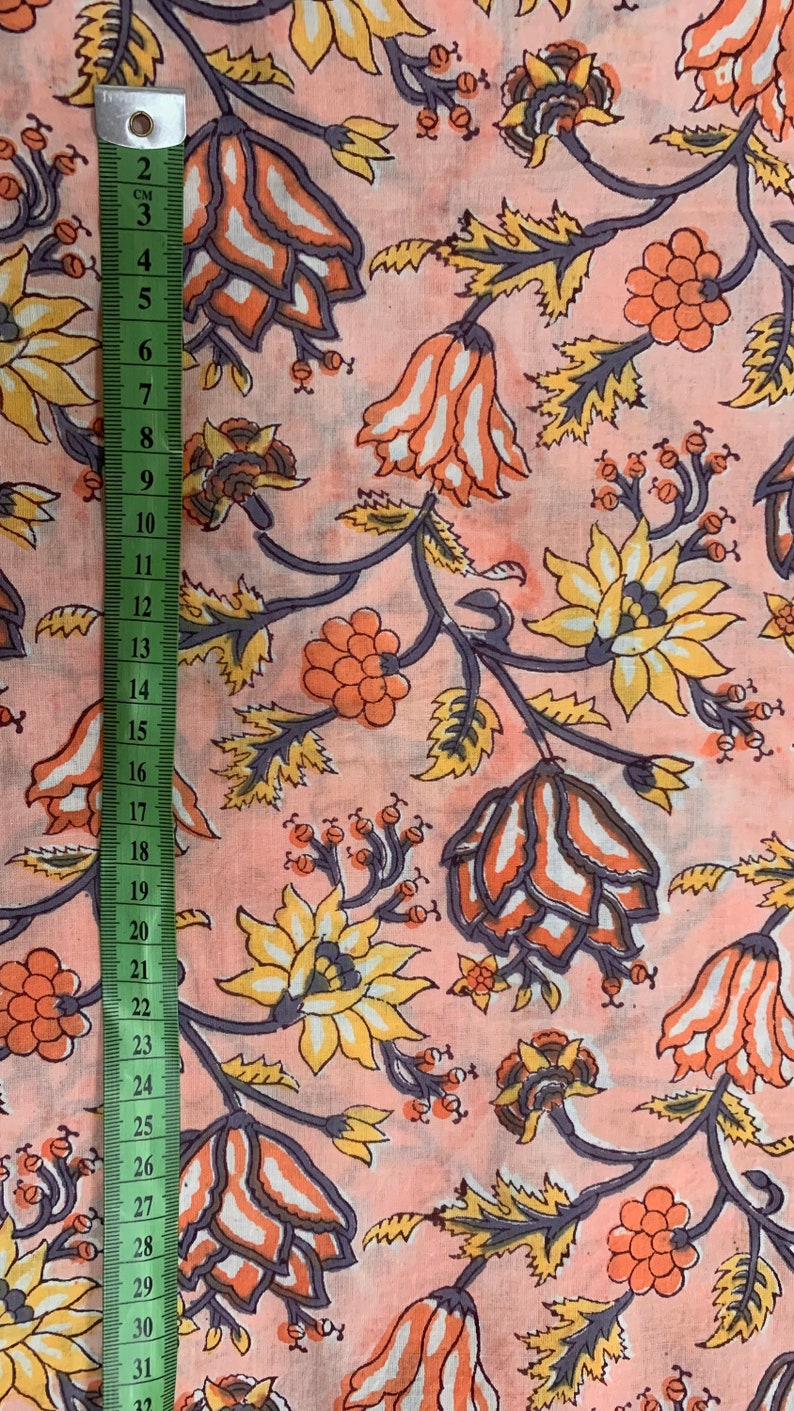 May include: Pink fabric with a floral print featuring orange, yellow, and white flowers with gray stems and leaves. The fabric is measured with a ruler in inches.