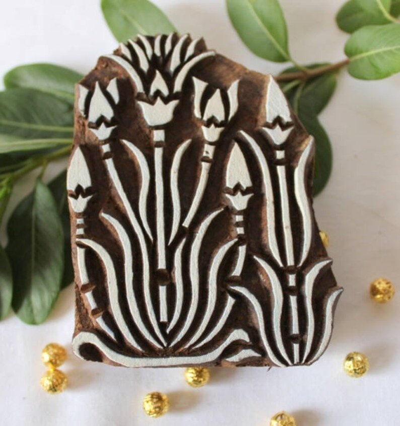May include: A wooden printing block with a floral design. The block is carved with a pattern of stylized flowers and leaves. The design is in white against a dark brown background.