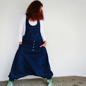 Può includere: Una donna indossa una tuta di jeans blu scuro con una canotta bianca sotto. La tuta ha una gamba larga e una vestibilità ampia. La donna è in piedi davanti a un muro bianco.