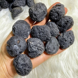 Puede incluir: Un puñado de piedras oscuras y redondas con una superficie texturizada. Las piedras son de color negro intenso y tienen una textura irregular. Las piedras se sostienen en una mano, con un fondo suave, blanco y esponjoso. Parecen ser piedras preciosas naturales y en bruto.