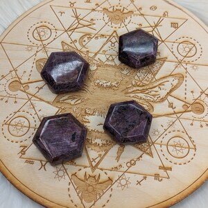 May include: Four faceted, hexagonal, dark purple crystals are arranged on a light wooden surface. The wooden surface is etched with geometric and celestial designs. The crystals have a polished, reflective surface.