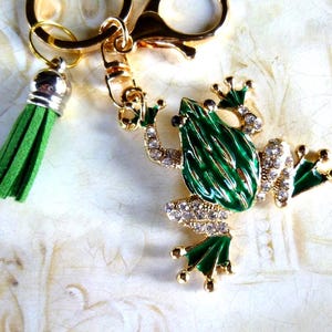 May include: A gold-coloured metal frog-shaped keyring with green enamel detailing and clear crystal accents. The frog has outstretched legs and a detailed design. A green tassel hangs from the keyring.