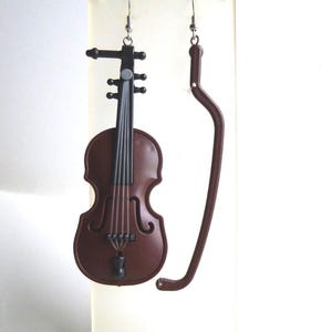 May include: Brown violin-shaped earrings with a matching brown bow. The earrings are suspended from silver-colored hooks. The violin has black strings and a black bridge. The bow is curved and has a rounded end.