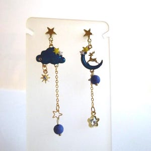 May include: Asymmetrical earrings featuring celestial designs. One earring has a dark blue cloud with stars, the other a crescent moon with a deer. Both have gold chains, stars, and blue spheres. A whimsical accessory.