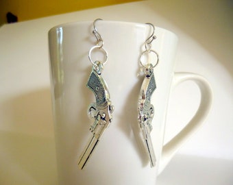May include: A pair of silver earrings with a gun charm. The earrings are hanging from a white mug.