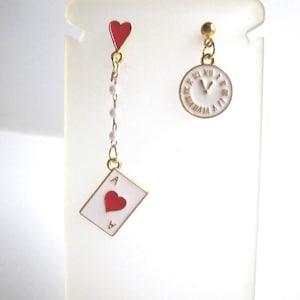 May include: Asymmetrical earrings with a playing card and clock theme. One earring features a white playing card with a red heart, and a red heart charm. The other earring is a white clock with gold Roman numerals.