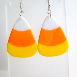 May include: A pair of candy corn earrings. Each earring is a triangular shape with white, orange, and yellow stripes. The earrings hang from silver-colored fish hook ear wires. The earrings are a festive accessory for Halloween.