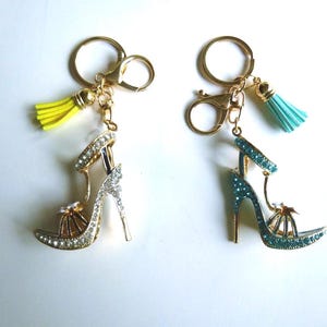 Könnte beinhalten: Zwei Schlüsselanhänger mit High-Heels-Schuhen. Einer ist silber- und goldfarben mit klaren Strasssteinen und einer gelben Quaste. Der andere ist türkis mit blauen Strasssteinen und einer türkisfarbenen Quaste. Beide haben goldfarbene Schlüsselringe.