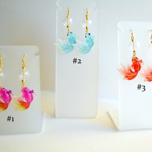 May include: Three pairs of dangle earrings featuring gold-tone hooks and clear glass beads. The earrings feature a goldfish design in pink and yellow, blue, and red.